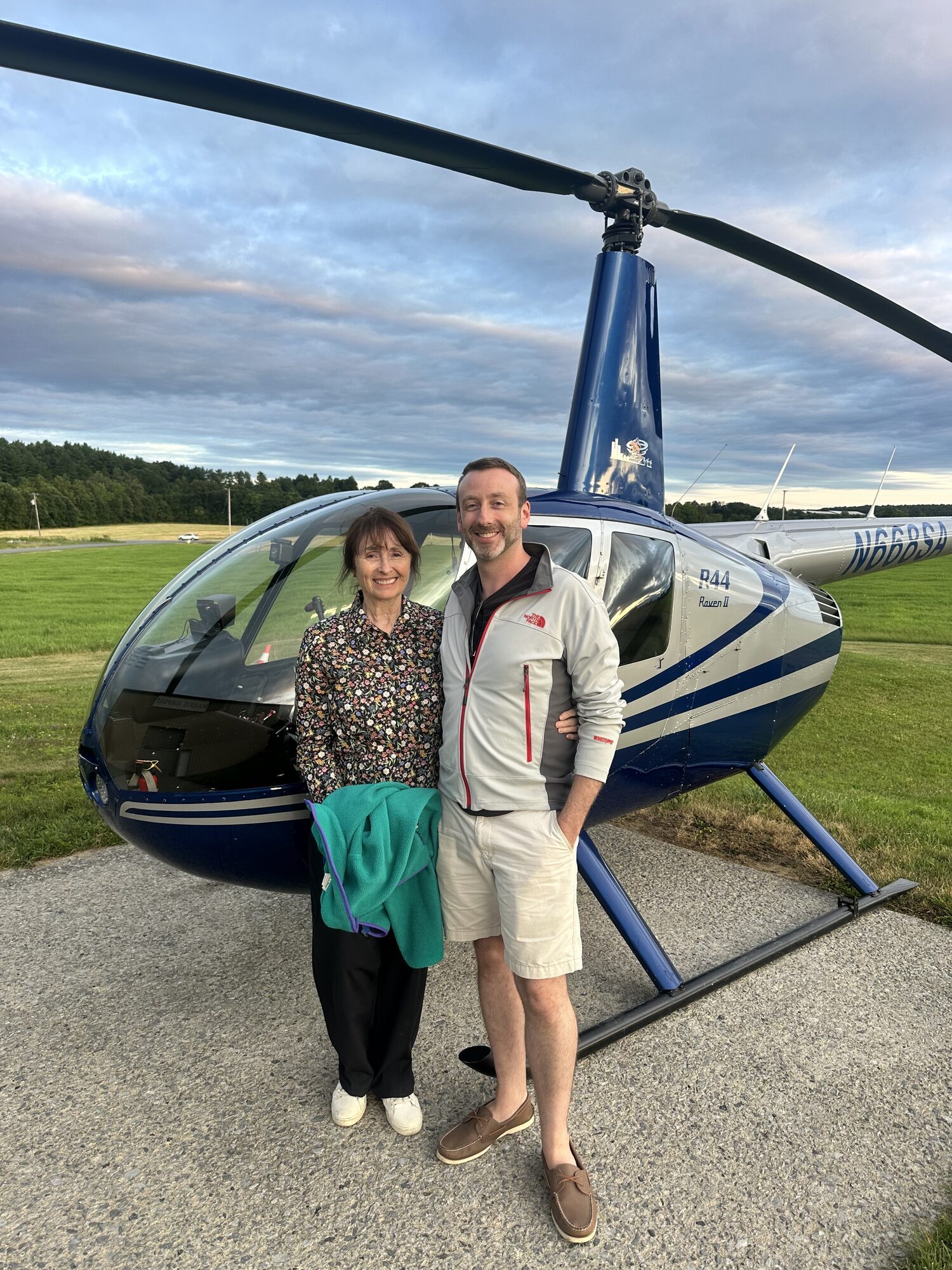 Two smiling guests beside the North Country Heli Flight Robinson R-44 Raven II after a Lake George tour at Harris Airport
